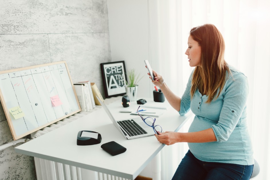 A pregnant young woman checks her mobile phone while working on a laptop sitting on her desk.