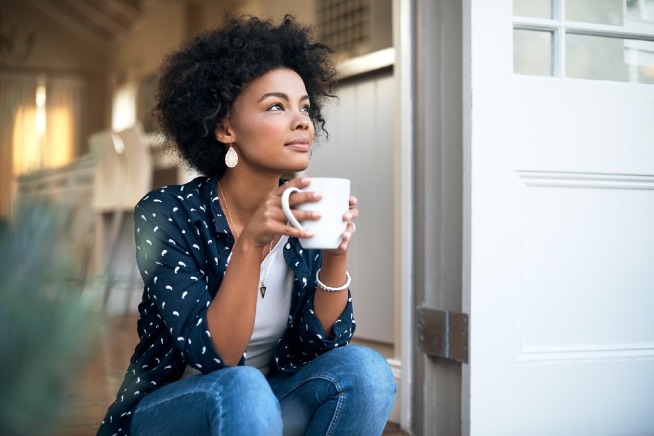 A young Black female sitting on her doorstep clasping a coffee mug and looking dreamily in the distance.