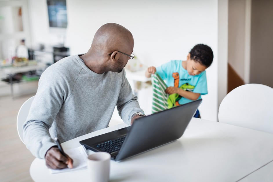 A man working from home on a laptop is distracted by his son folding a towel in the background