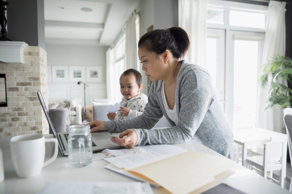 A young asian mother making an isnurance payment on a laptop while her baby sits on the table next to her