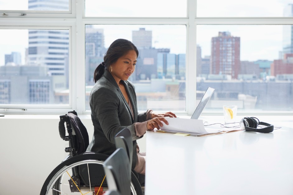 A disabled black female on a wheelchair sitting at a table looking through some papers and a laptop. City high rises visible in the background window.