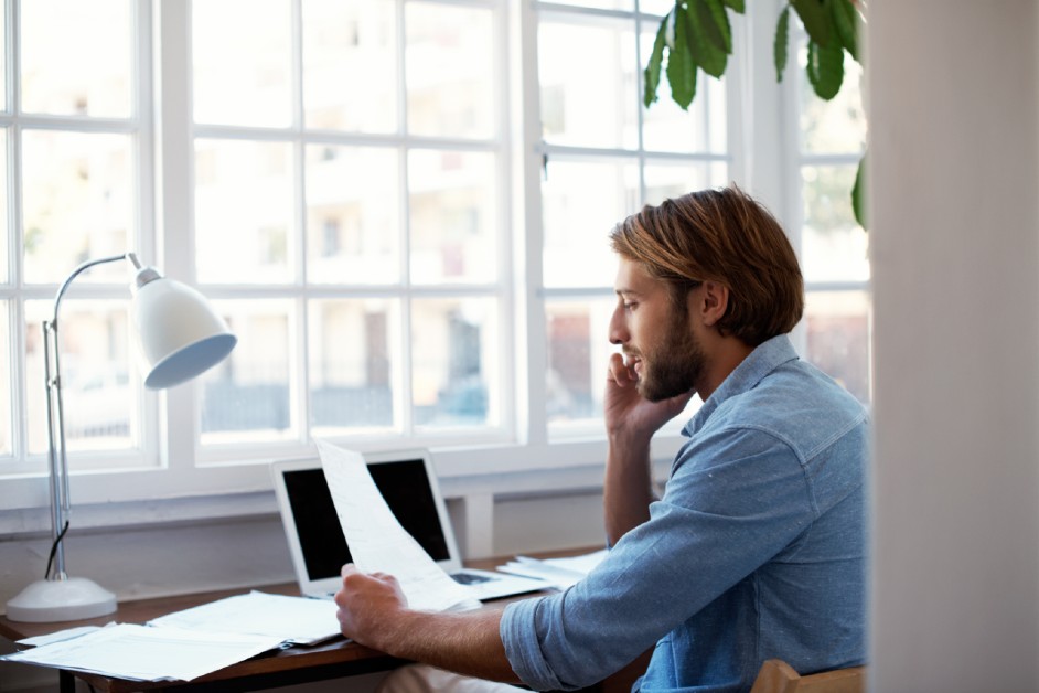 A man sitting on his work desk, holding some paperwork, and talking on the phone. More paperwork, a laptop and table lamp are seen on the desk.