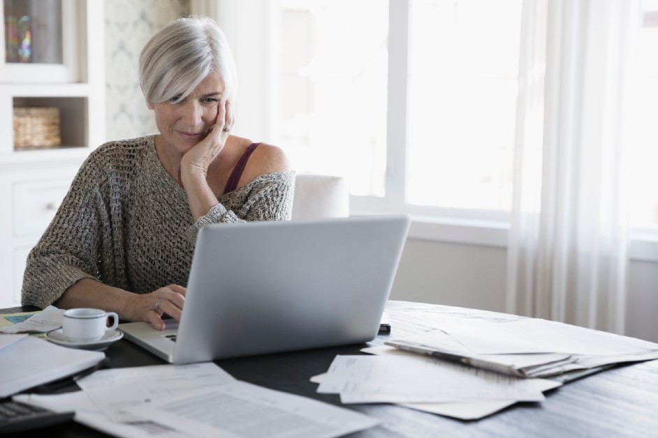 A middle-aged lady reading something on her laptop on a table with lots of paperwork strewn around and a cup of tea beside her.
