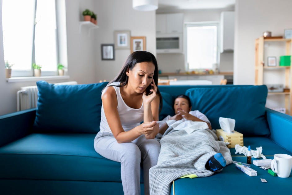 A young mom checking her child's temperature and making a call while the kid lies on the couch visibly sick. Medicine and tissue papers strewn around.
