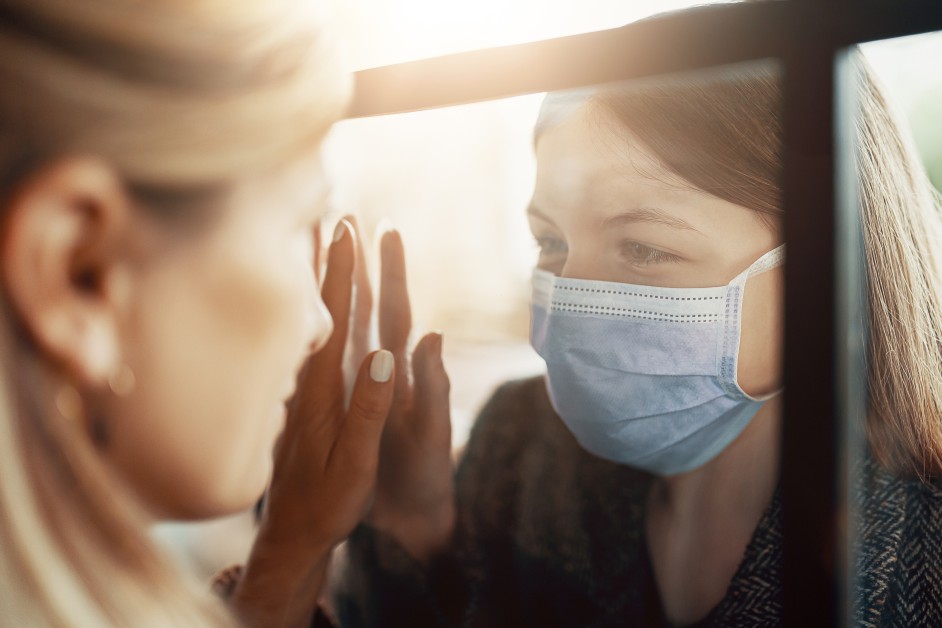 Two women meeting each other through a glass wall, one wearing a mask.